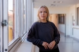A woman with blonde hair and a dark sweater poses for a photo by a window in a hospital corridor