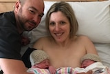 A woman in a hospital bed holding newborn twins with her husband by her side