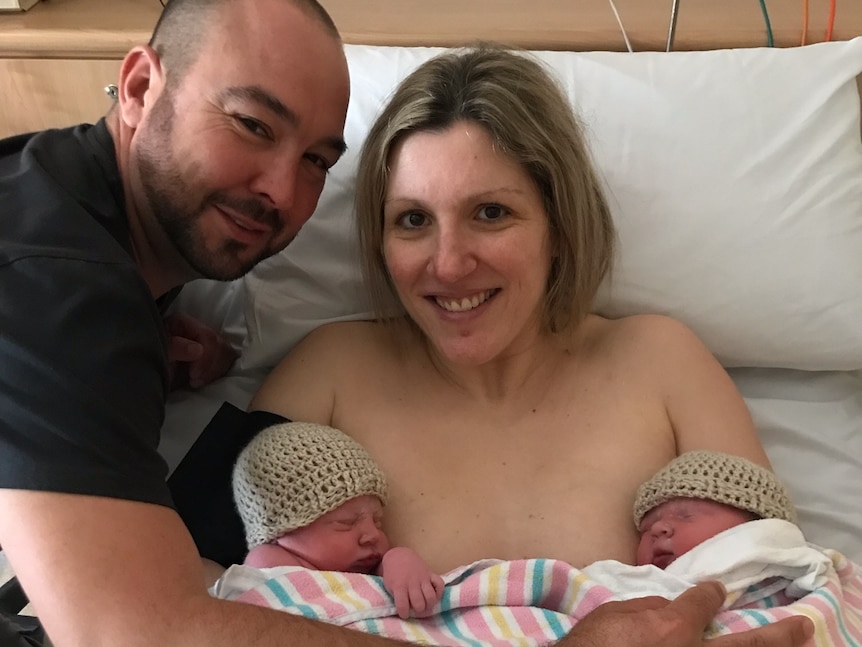 A woman in a hospital bed holding newborn twins with her husband by her side