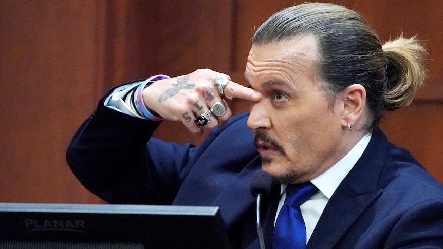 Johnny Depp in the witness stand in a court pointing at his own face. 