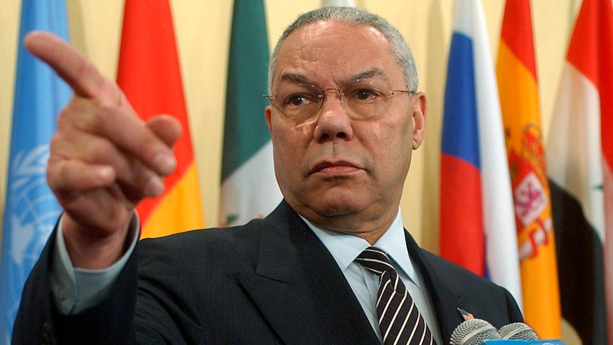Colin Powell, standing in front of flags, points to a reporter during a news conference.
