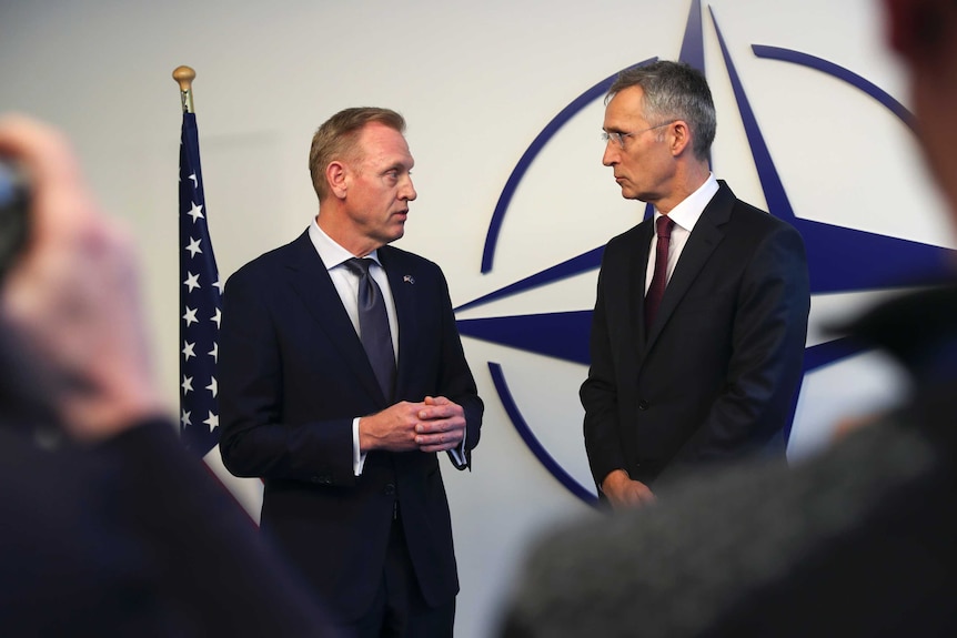 Two men in navy blue suits talk in front of the US flag and the NATO logo, a blue four-spoke star pointing north.