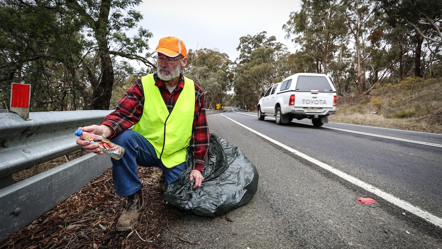 Bill Wiglesworth crouched on the side of the Pyrenees Highway holding a discarded plastic bottle and garbage bag.