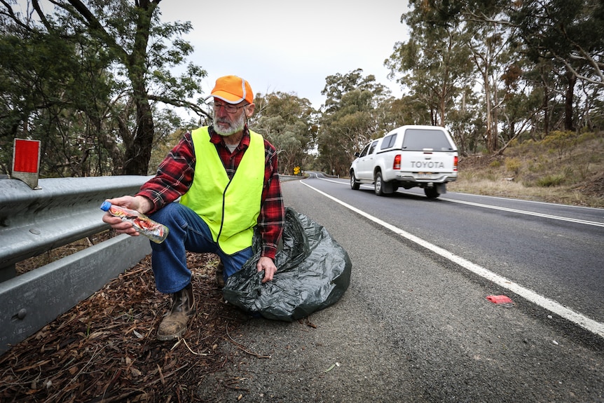 Bill Wiglesworth crouched on the side of the Pyrenees Highway holding a discarded plastic bottle and garbage bag.