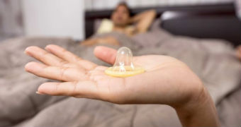 A hand holding a condom near a bed.