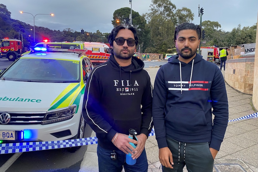 two men in dark clothing, one wearing sunglasses, in front of a crash site and ambulance vehicle