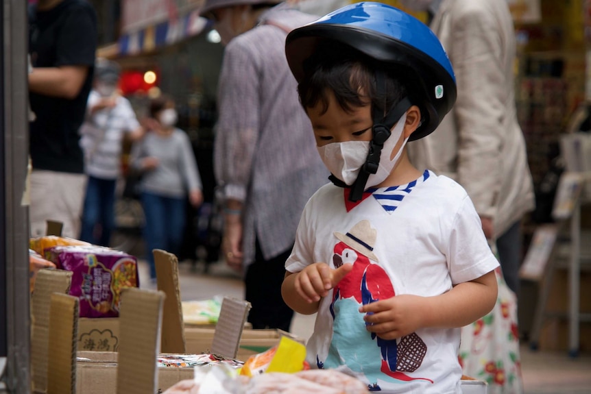 A child in a face mask and bike helmet examines food at a stall in Tokyo