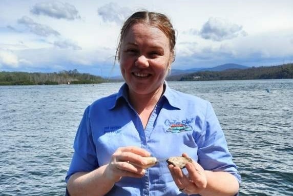A woman stands holding an oyster, wearing a blue shirt with water in the background