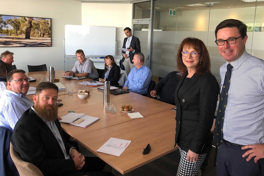 A group of people sit around a boardroom table; in the foreground stand a man and a woman, both wearing glasses.