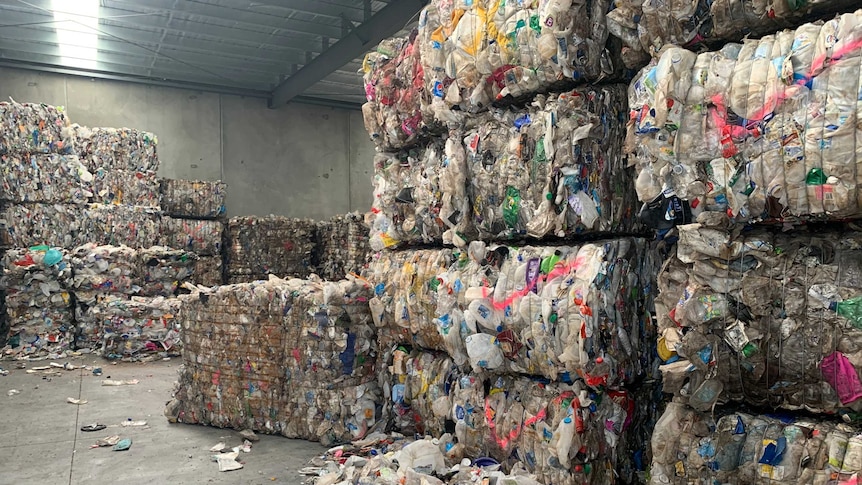 A picture of rubbish packaged into squares and piled all through a grey warehouse. Loose rubbish can be seen on the ground.