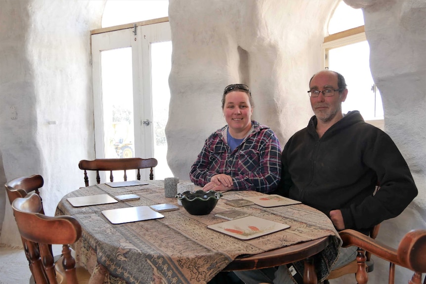 A man and woman sit at a table inside a white room with windows and doors.