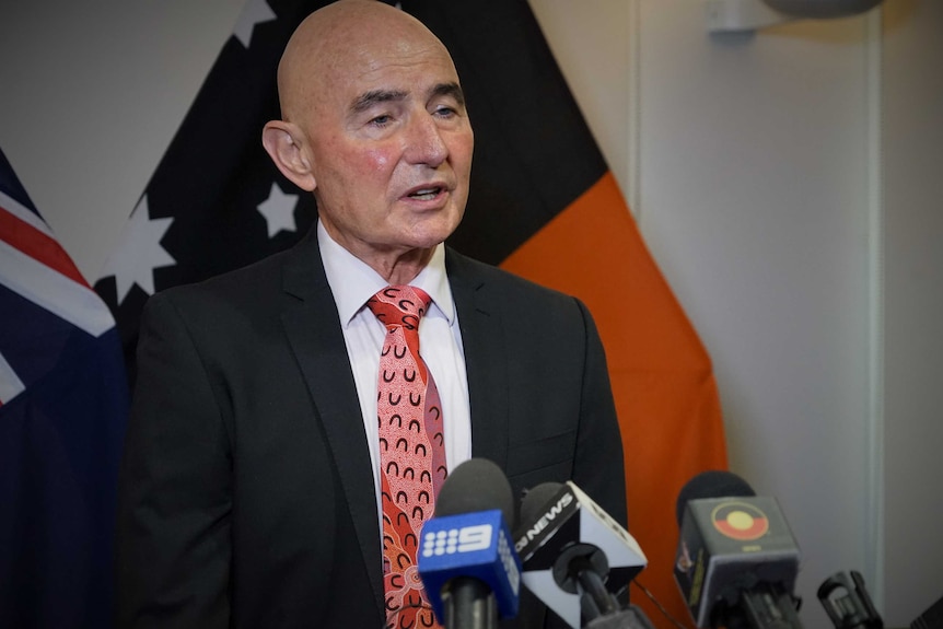 Hugh Heggie is wearing a suit and tie and standing in front of a NT flag. He is talking at a press conference.