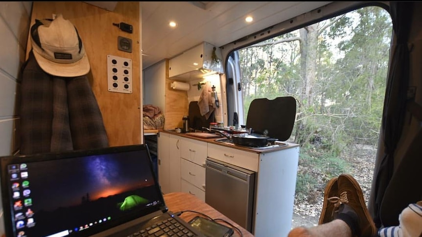 A man relaxes in a campervan with his laptop by his side. The bush can be seen from the window.