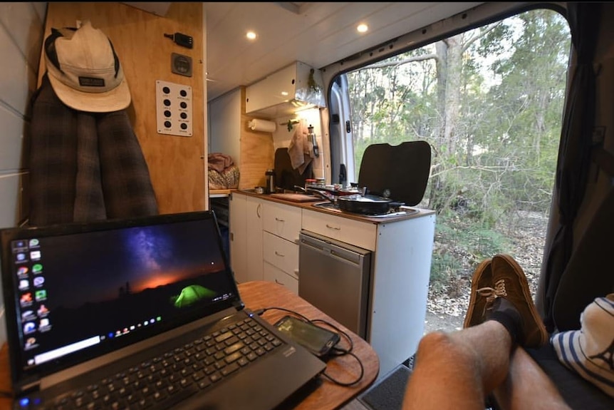 A man relaxes in a campervan with his laptop by his side. The bush can be seen from the window.