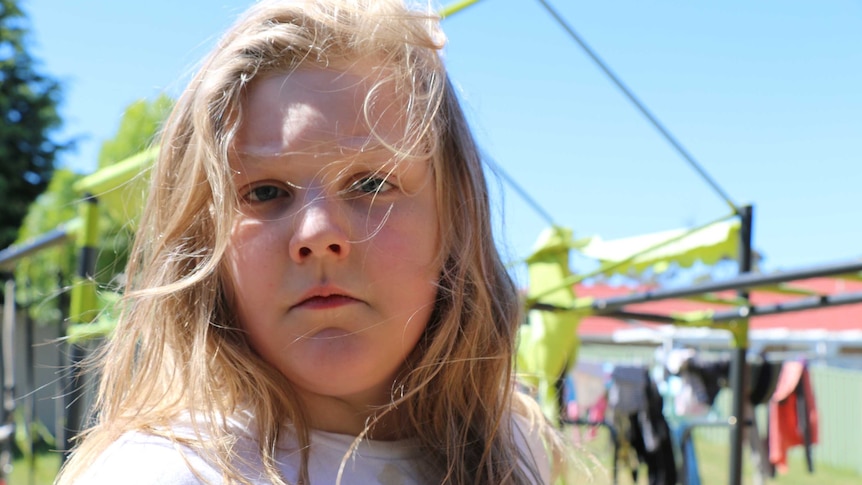 A young boy with long blonde hair stands in a backyard.