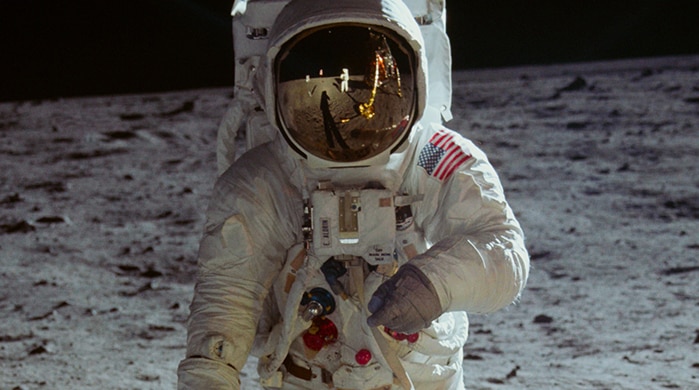 An astronaut standing on the moon. The reflection of their spacesuit's gold visor shows another astronaut and space craft.