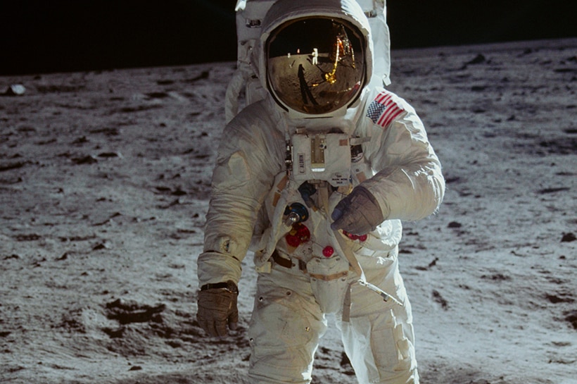 An astronaut standing on the moon. The reflection of their spacesuit's gold visor shows another astronaut and space craft.