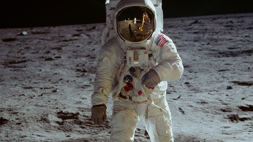 An astronaut standing on the moon. The reflection of their spacesuit's gold visor shows another astronaut and space craft.