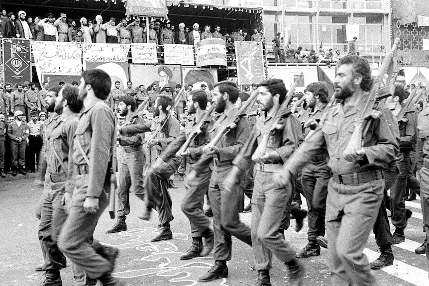 A black and white photo shows rows of soldiers carrying rifles parading past an official viewing stand.