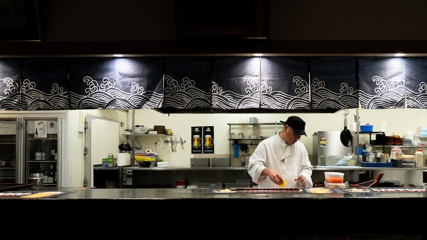 A lone chef in an open kitchen, framed by a silhouette of the kitchen wall.