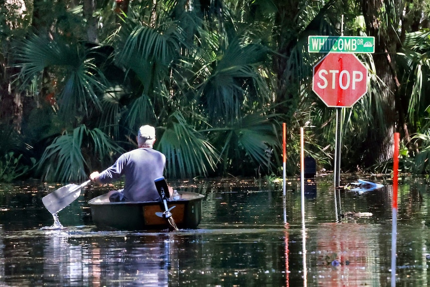 A man paddles in a canoe down a flooded street news a stop sign. 
