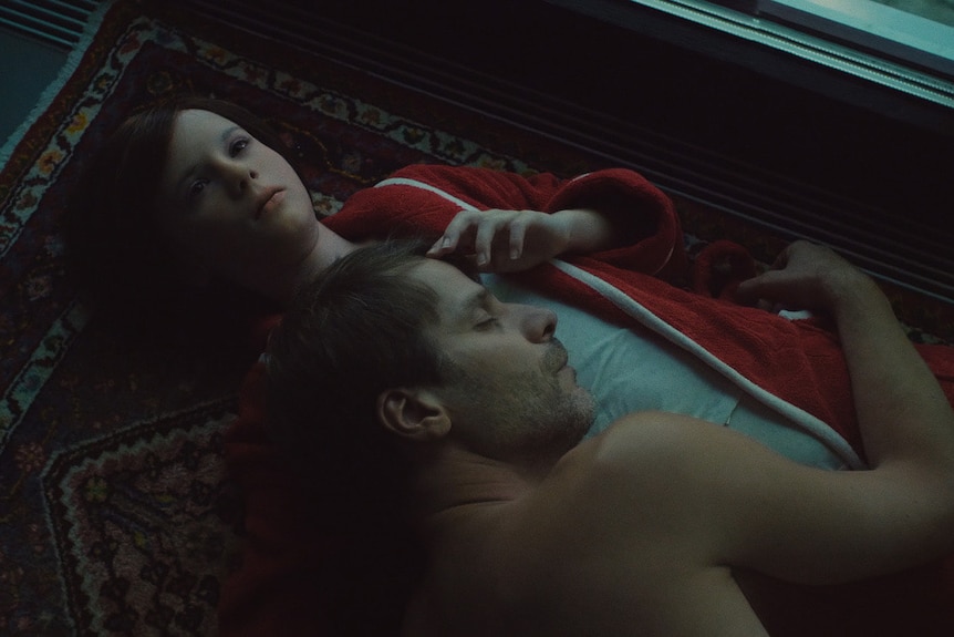 A man lies in the arms of a young AI girl in the film The Trouble with Being Born