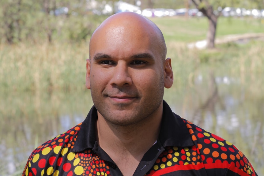 Indigenous man wearing a shirt with traditional art in red, orange and yellow.