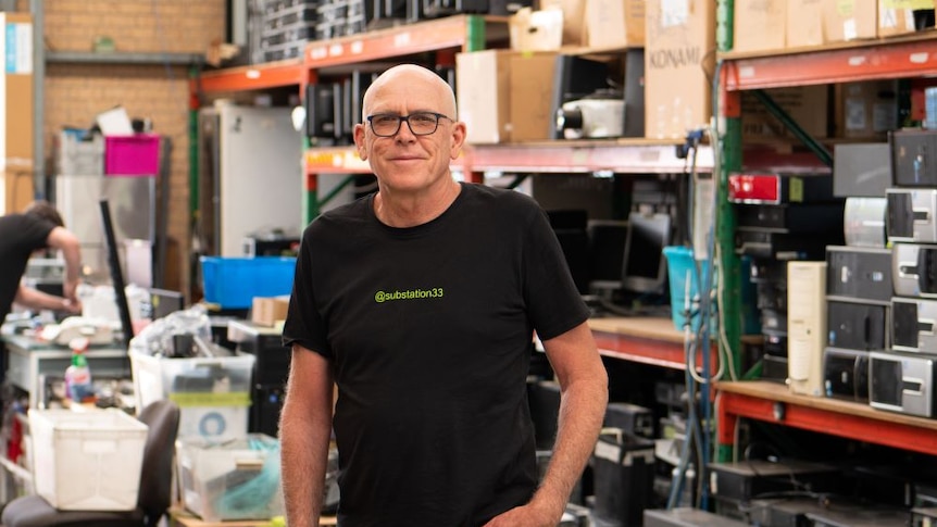 A bald man stands smiling in front of many plastic containers. He wears a black Substation 33 shirt.