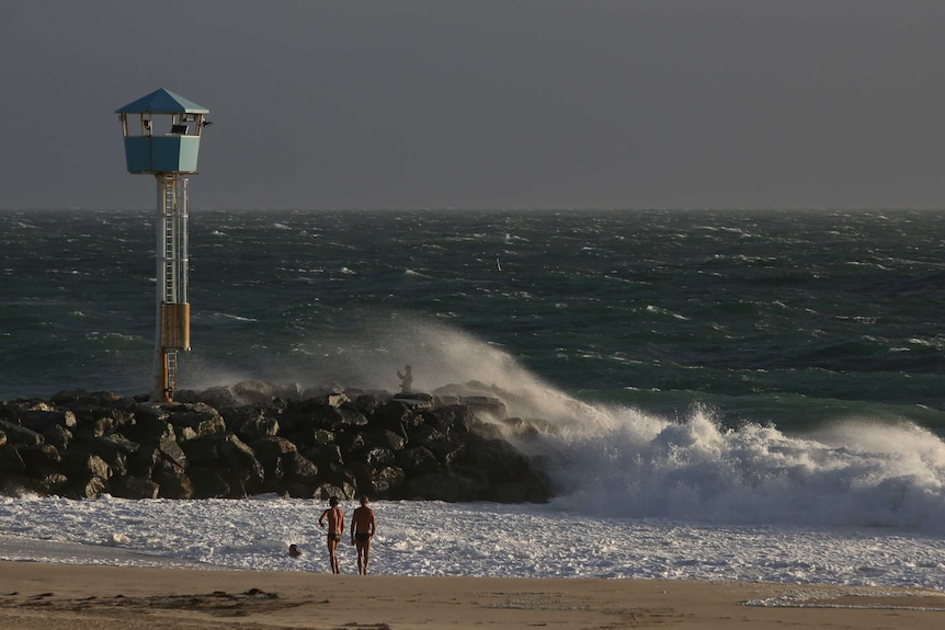 A wave crashes over a person on rocks at City Beach and two boys approach the water.
