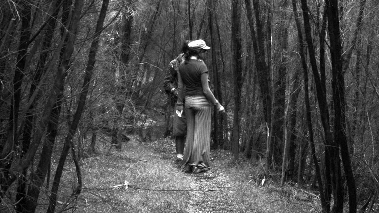 A black and white photograph of a woman and man behind her in a grove of trees on a bush track