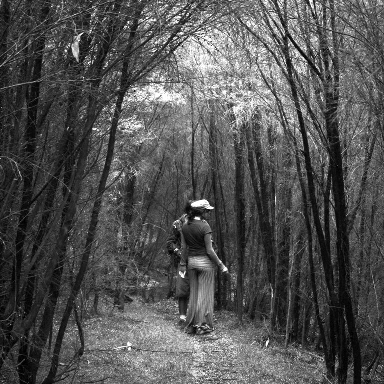 A black and white photograph of a woman and man behind her in a grove of trees on a bush track