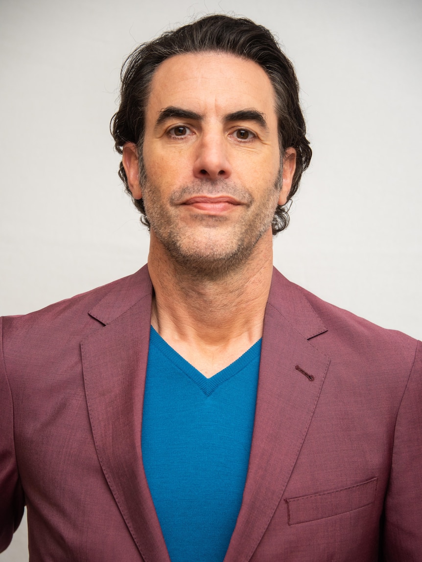 A head shot of Sacha Baron Cohen smiling slightly wearing a maroon suit jacket and blue shirt