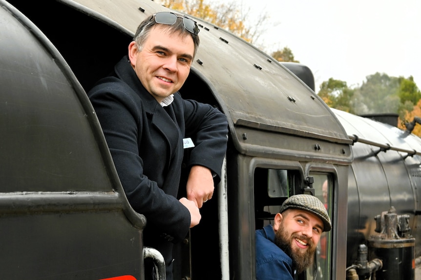 A man leans out of the cab of a steam train engine with another looking up at him