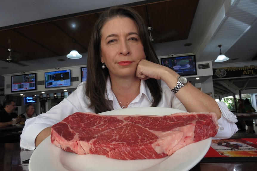 A woman with long dark hair sits and stares into the camera above a massive piece of steak 