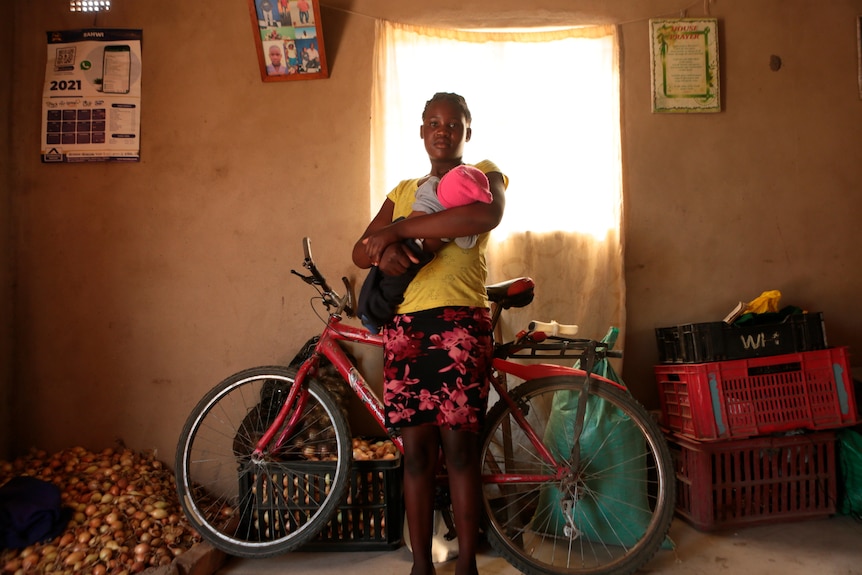 A young girl holding an infant stands in front of a bicycle next to a window with a pile of onions in the background.