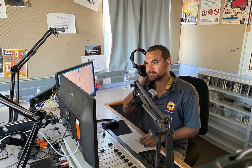 An Aboriginal man with a beard and short hair holds a headphone to his right ear in a radio studio.