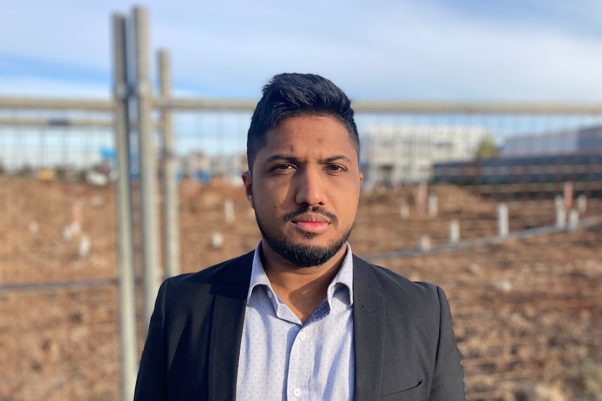 Jazeer Nijamudeen wears a suit jacket and stands looking seriously in front of a fence and soil.
