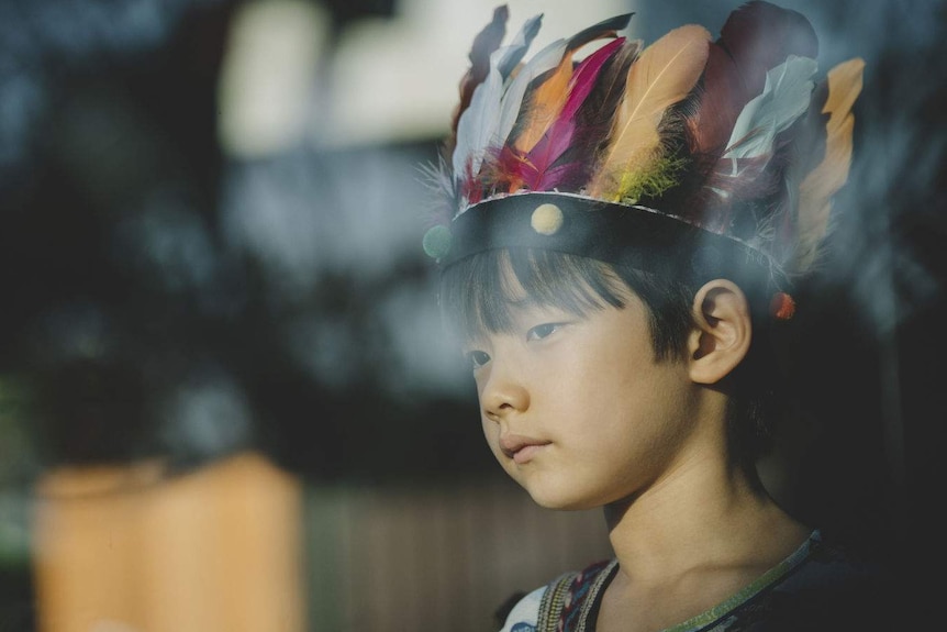 A small boy wearing a feathered headdress stares through a lightly reflective window.