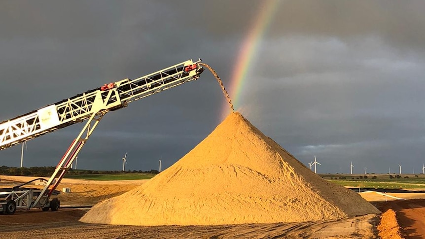 A rainbow ends behind a pile of yellow sand, as more sand is added by a machine. The sky is dark and stormy.