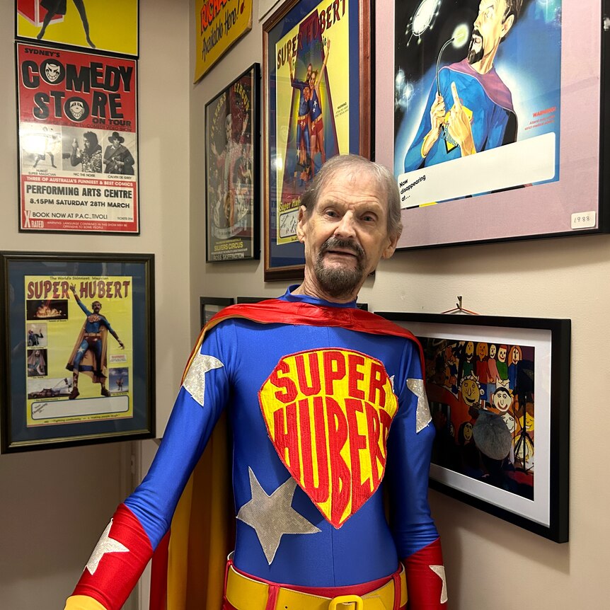 A man wearing a blue and red super suit stands in a hallway with photos in frames on the wall.