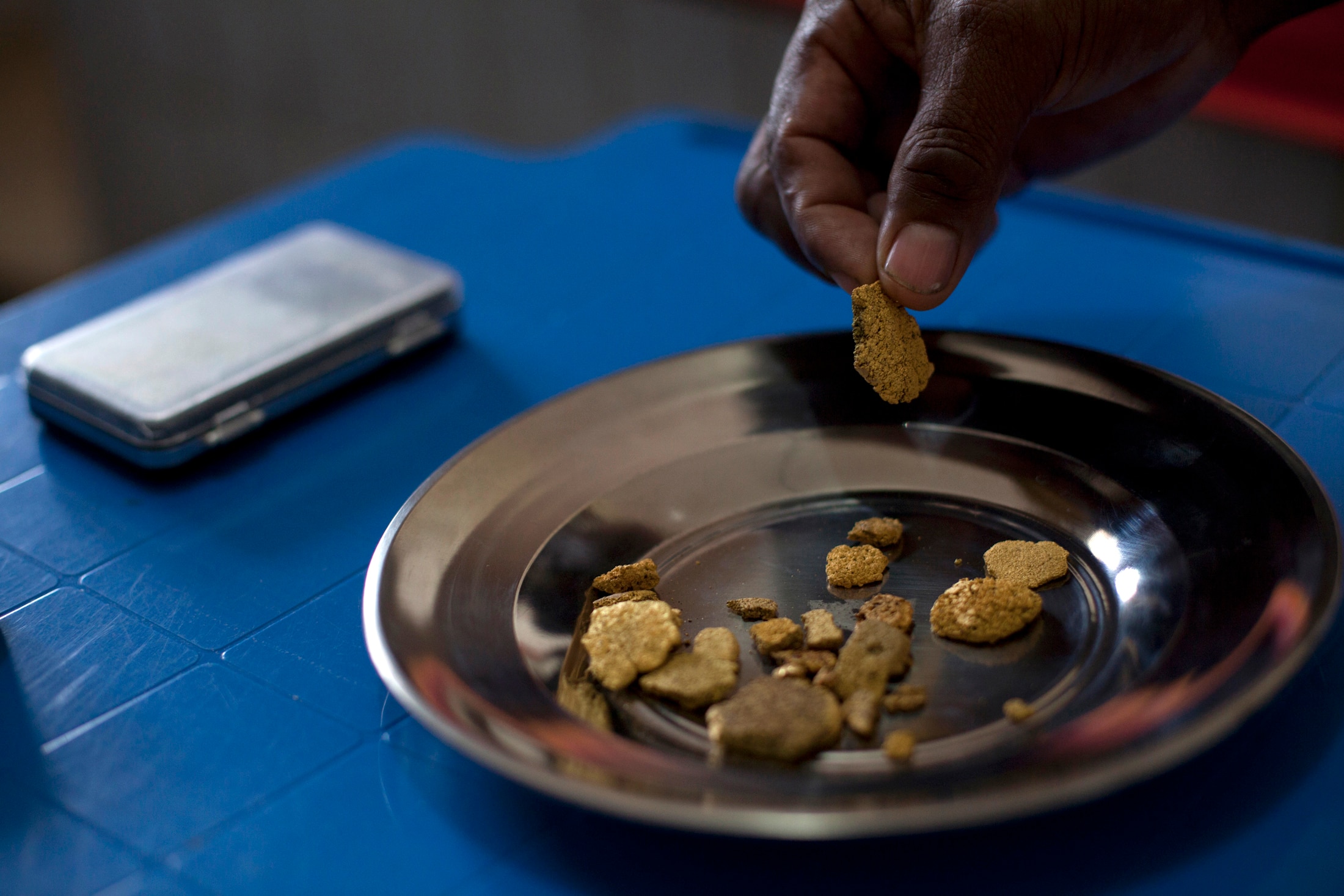 A person's hand holding a small chunk of gold above a silver plate with other gold chips, on a blue table.