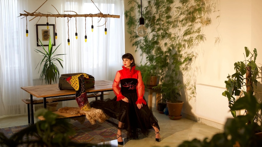 An Afghan woman in red and black dress sits in an apartment filled with plants and beautiful objects