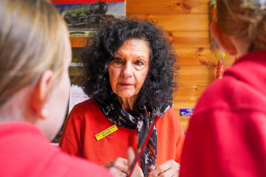 An older woman with curly black hair in a red blazer and name tag talks to two young girls in school uniform.