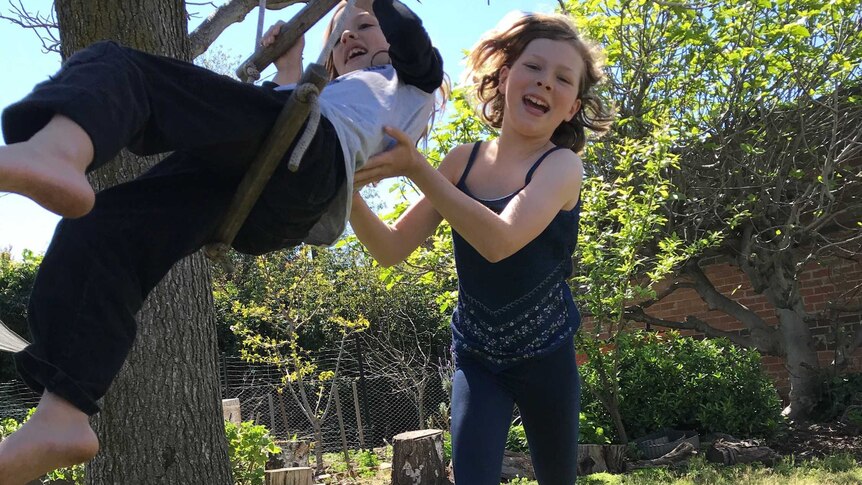 A boy sits on a rope ladder being pushed by a girl in a backyard setting.