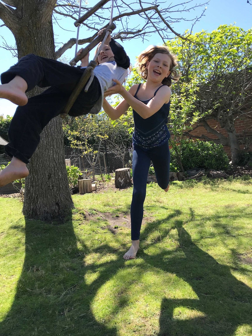 A boy sits on a rope ladder being pushed by a girl in a backyard setting.