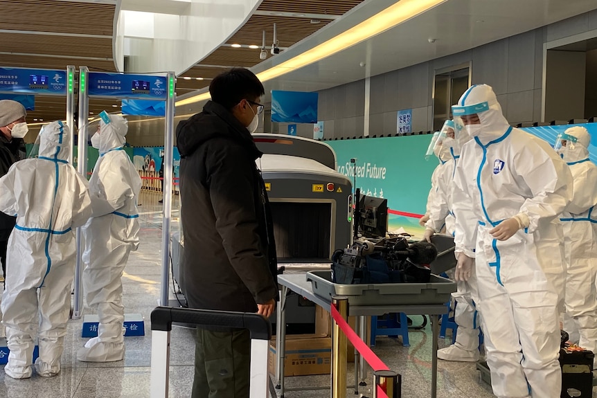 A man waits at airport security as his bags are scanned by teams wearing full PPE and face shields.