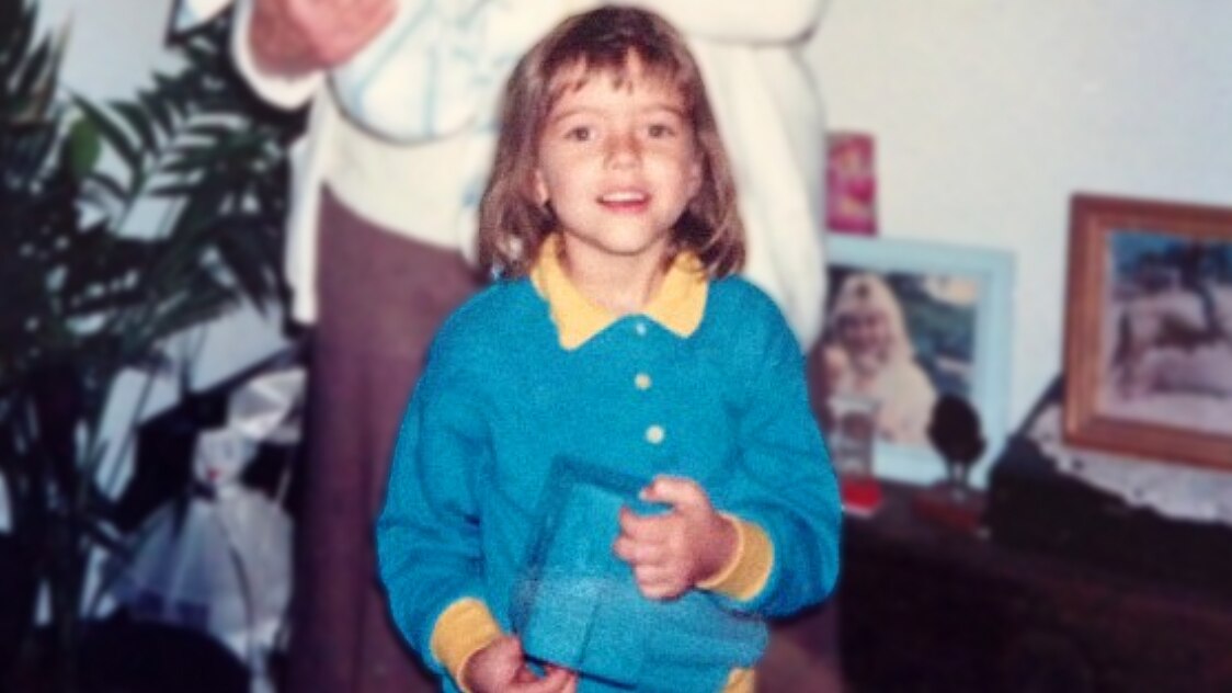An old photo of a smiling 5-year-old girl in a turquoise cardigan with a yellow collar and sleeves. She is holding a blue box.