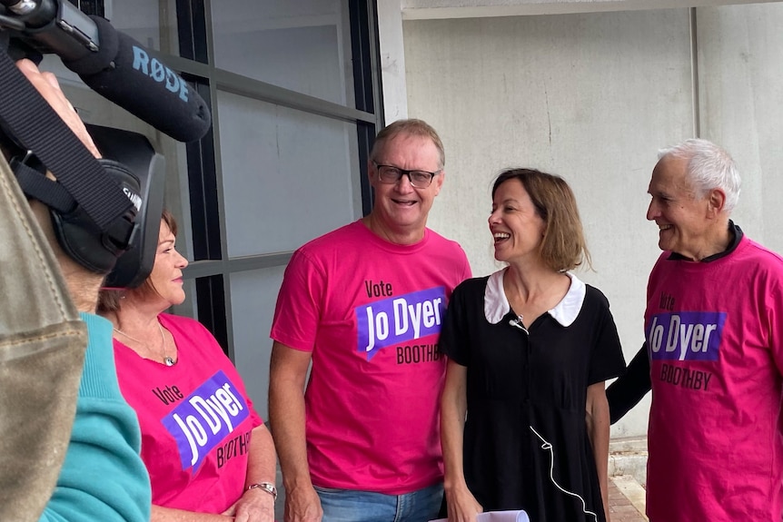 A woman wearing white collar, black top, stands in between two men in pink 'Jo Dyer' logo t-shirts.