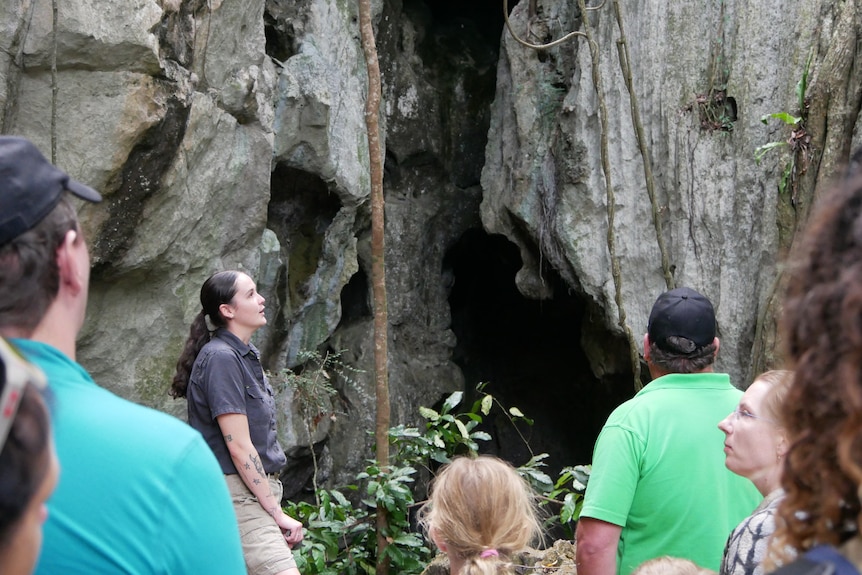 A group of people admire a large cave with a woman who is their guide.