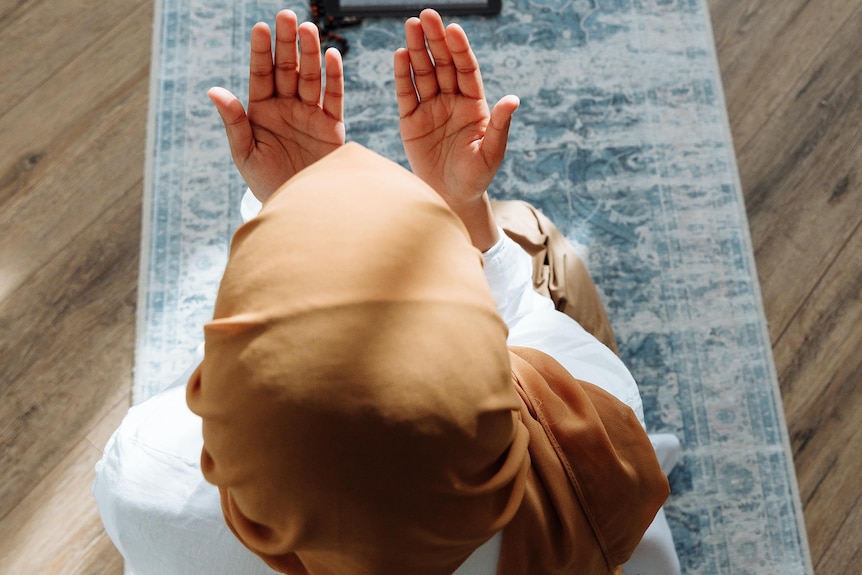 A person is sitting on a prayer mat with their hands unfolded.